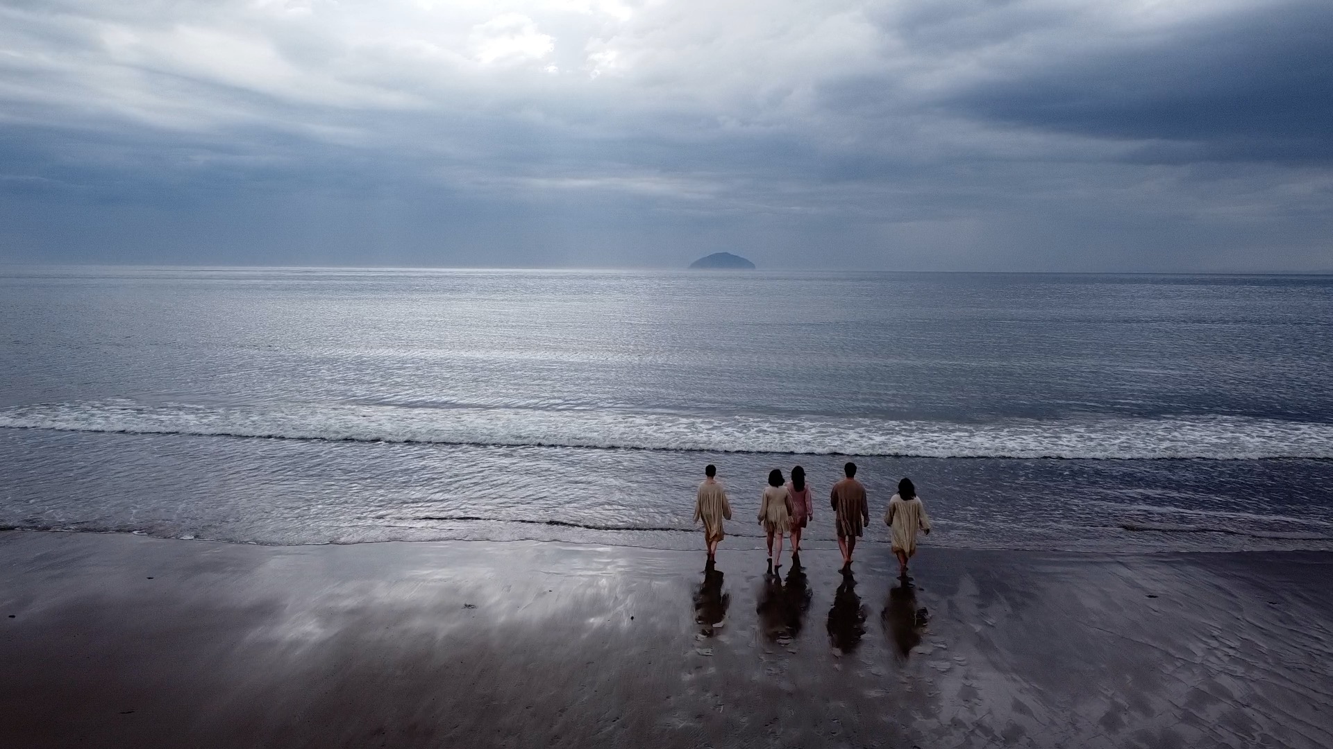 An arial shot of a beach and sea, as 5 people in cream coloured smocks walk into the water. It has an eerie feel.
